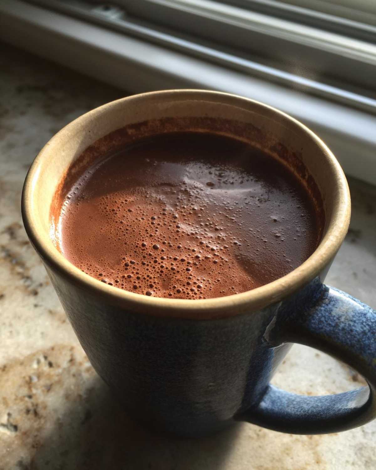 Close-up of a steaming mug filled with rich, dark hot chocolate, sitting near a window.