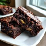 Two fudgy, rich squares of tasty healthy date brownies stacked on a white plate, showing visible walnut pieces.