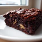 A rich, dark chocolate square of tasty healthy date brownies featuring visible pieces of walnuts on a white plate.