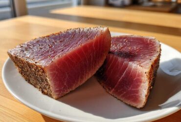 Two thick, rare tuna steaks with a pepper crust resting on a white plate, ready for a cast iron skillet sear.