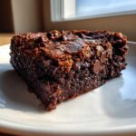 A close-up, rich, dark slice of Ultimate Fudgy Zucchini Brownies with visible chocolate chips on a white plate.