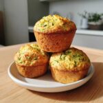 Three golden-brown Ultimate Moist Zucchini Muffins stacked on a small white plate, showing visible shreds of green zucchini.