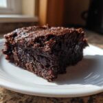 A close-up of a rich, fudgy slice of Vegan Avocado Zucchini Brownies on a white plate.