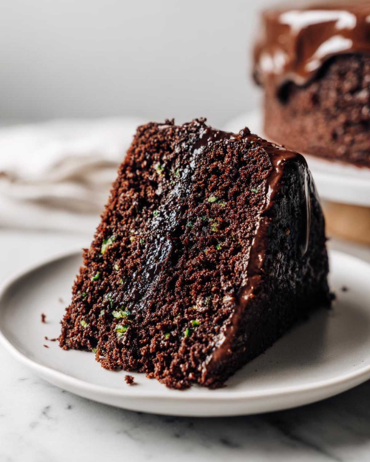 A moist slice of rich chocolate cake showing flecks of green zucchini, topped with glossy chocolate ganache. This is the Vegan Chocolate Zucchini Cake.
