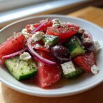 Close-up of a fresh Greek salad featuring chunks of tomato, cucumber, red onion, feta cheese, and Kalamata olives, seasoned with herbs.