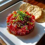 A vibrant mound of diced red tuna in a Tuna Tartare Recipe, topped with sesame seeds and scallions, served with crackers.