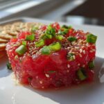 Close-up of bright red, cubed Tuna Tartare Recipe garnished with sesame seeds and green onions, served next to crackers.
