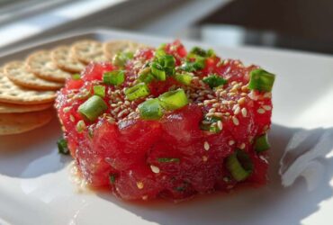 Close-up of bright red, cubed Tuna Tartare Recipe garnished with sesame seeds and green onions, served next to crackers.