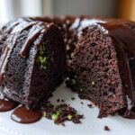 A rich, dark chocolate bundt cake, cut open to show green zucchini shreds inside, covered in dripping chocolate glaze.