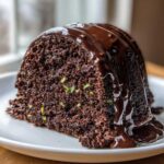 A close-up of a slice of rich, dark chocolate zucchini bundt cake topped with dripping chocolate glaze.