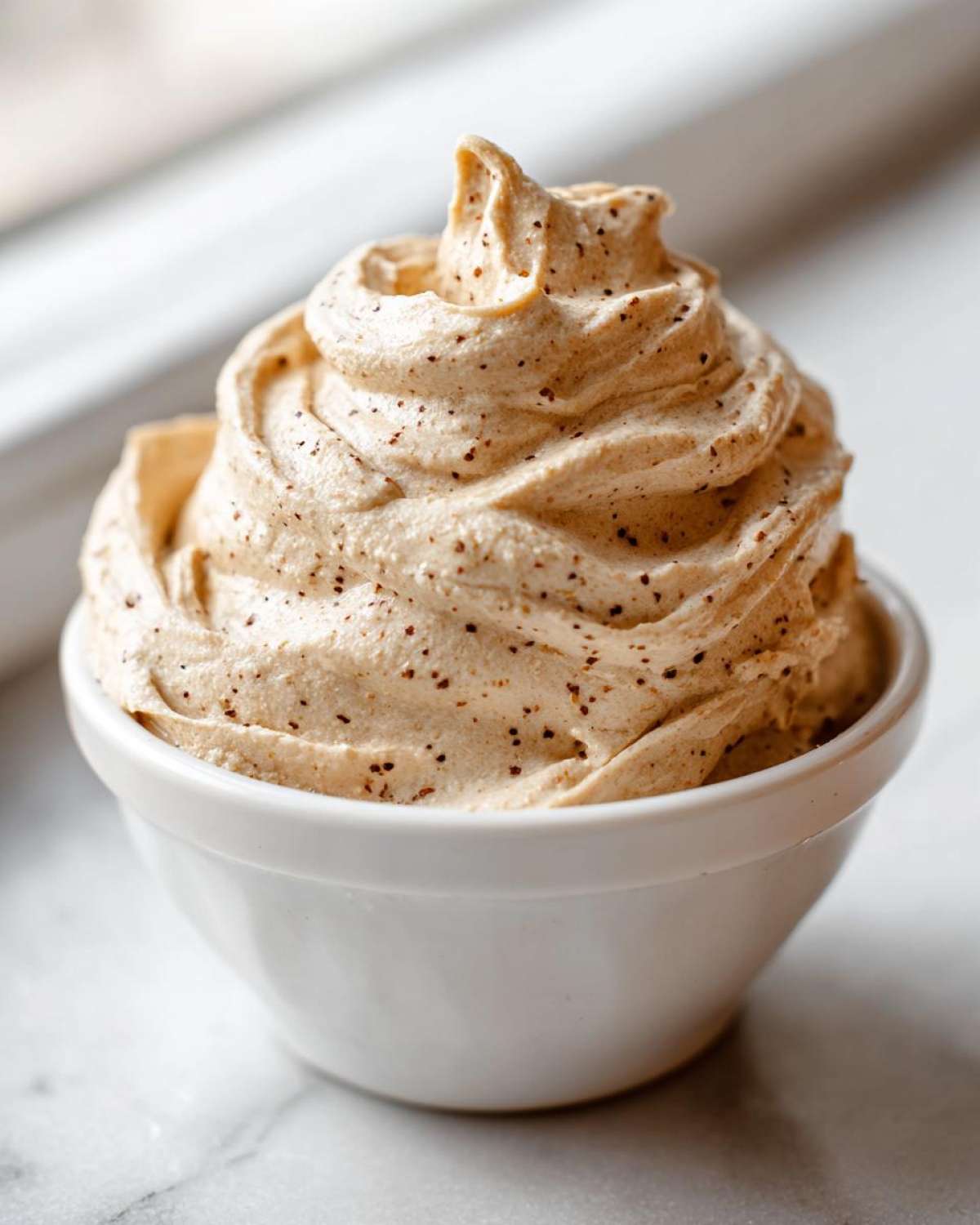 A close-up of fluffy, whipped cinnamon butter piped into a small white bowl, showing dark spice specks throughout.