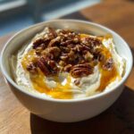 A white bowl filled with creamy whipped goat cheese, drizzled with honey and topped generously with toasted pecans.
