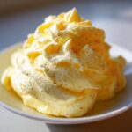 Close-up of a large swirl of light yellow, whipped honey butter, speckled with tiny brown flecks, served on a small white dish.