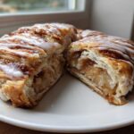 A cross-section view of a flaky Wisconsin apple kringle recipe pastry, filled with spiced apples and topped with white icing.