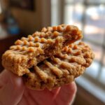 A hand holding a peanut butter cookie broken in half, showing the soft, crumbly interior texture of the peanut butter cookies.