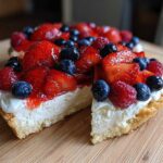 A close-up slice cut from an Amazing Berry Fruit Pizza showing a cookie crust, cream cheese filling, and topping of strawberries, blueberries, and raspberries.