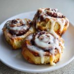 Three freshly baked Banana Bread Batter Cinnamon Buns generously drizzled with white icing on a white plate.