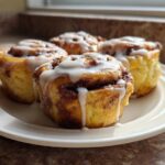 Four freshly baked Banana Bread Batter Cinnamon Buns drizzled with white icing on a white plate.