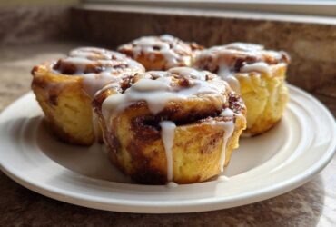 Four freshly baked Banana Bread Batter Cinnamon Buns drizzled with white icing on a white plate.
