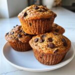 A stack of four freshly baked Banana Chocolate Chip Muffins topped with melted chocolate chips on a white plate.