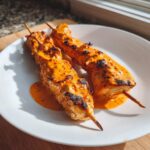 Two grilled Bang Bang Chicken Skewers generously coated in a creamy, spicy orange sauce, resting on a white plate.