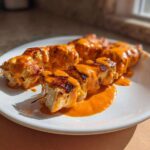 Close-up of grilled Bang Bang Chicken Skewers generously drizzled with a creamy, bright orange sauce on a white plate.