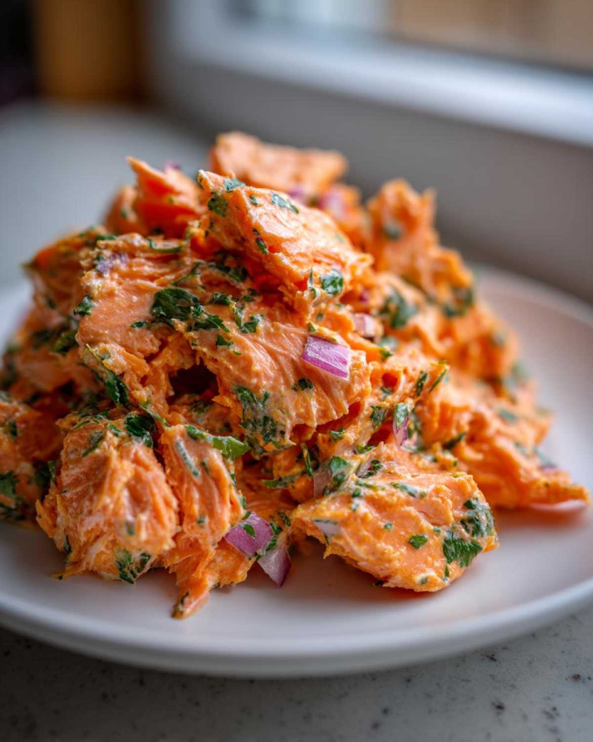 A close-up of creamy, flaky Bang Bang Salmon Salad mixed with chopped herbs and red onion pieces on a white plate.