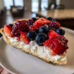 A close-up slice of Berry Fruit Pizza showing a thick crust, white cream cheese layer, and glossy mixed berries.