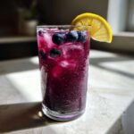 A tall, frosty glass filled with vibrant purple Blueberry Lemonade, ice cubes, fresh blueberries, and a lemon slice garnish.