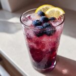 A tall glass filled with vibrant Blueberry Lemonade, ice cubes, fresh blueberries, and lemon slices.