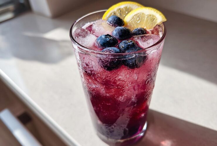 A tall glass filled with vibrant Blueberry Lemonade, ice cubes, fresh blueberries, and lemon slices.