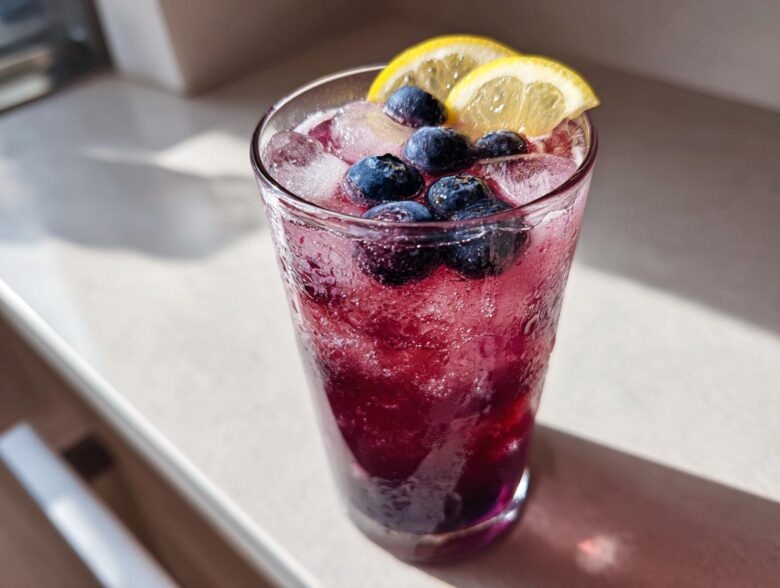 A tall glass filled with vibrant Blueberry Lemonade, ice cubes, fresh blueberries, and lemon slices.