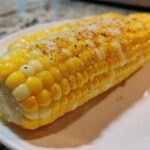 Close-up of two ears of cooked Buttered Corn on the cob glistening with butter and topped with salt and pepper.
