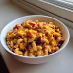 A white bowl filled with cheesy Vegetarian Chili Mac, featuring elbow macaroni, kidney beans, and melted cheddar cheese.
