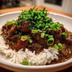 A close-up of rich, dark Chicken Étouffée served over fluffy white rice, heavily garnished with fresh parsley and green onions.