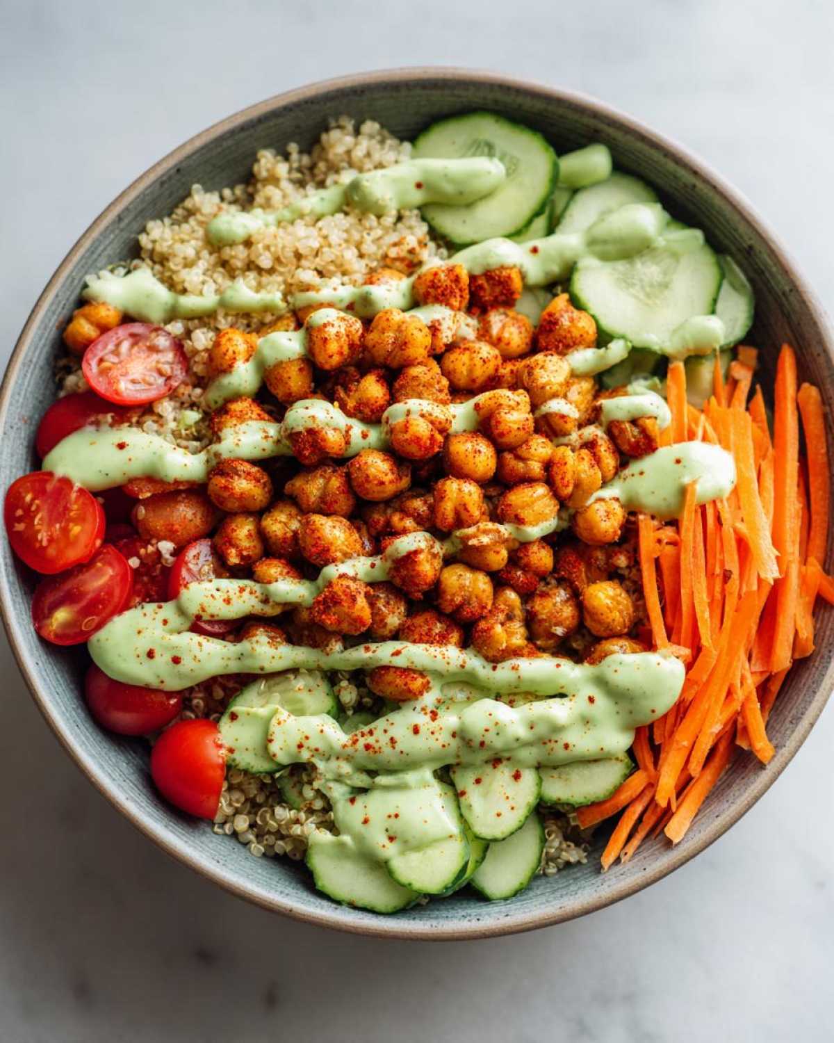 Overhead view of a vibrant Chickpea Grain Bowl with avocado lemon tahini dressing, quinoa, roasted chickpeas, cucumbers, carrots, and tomatoes.