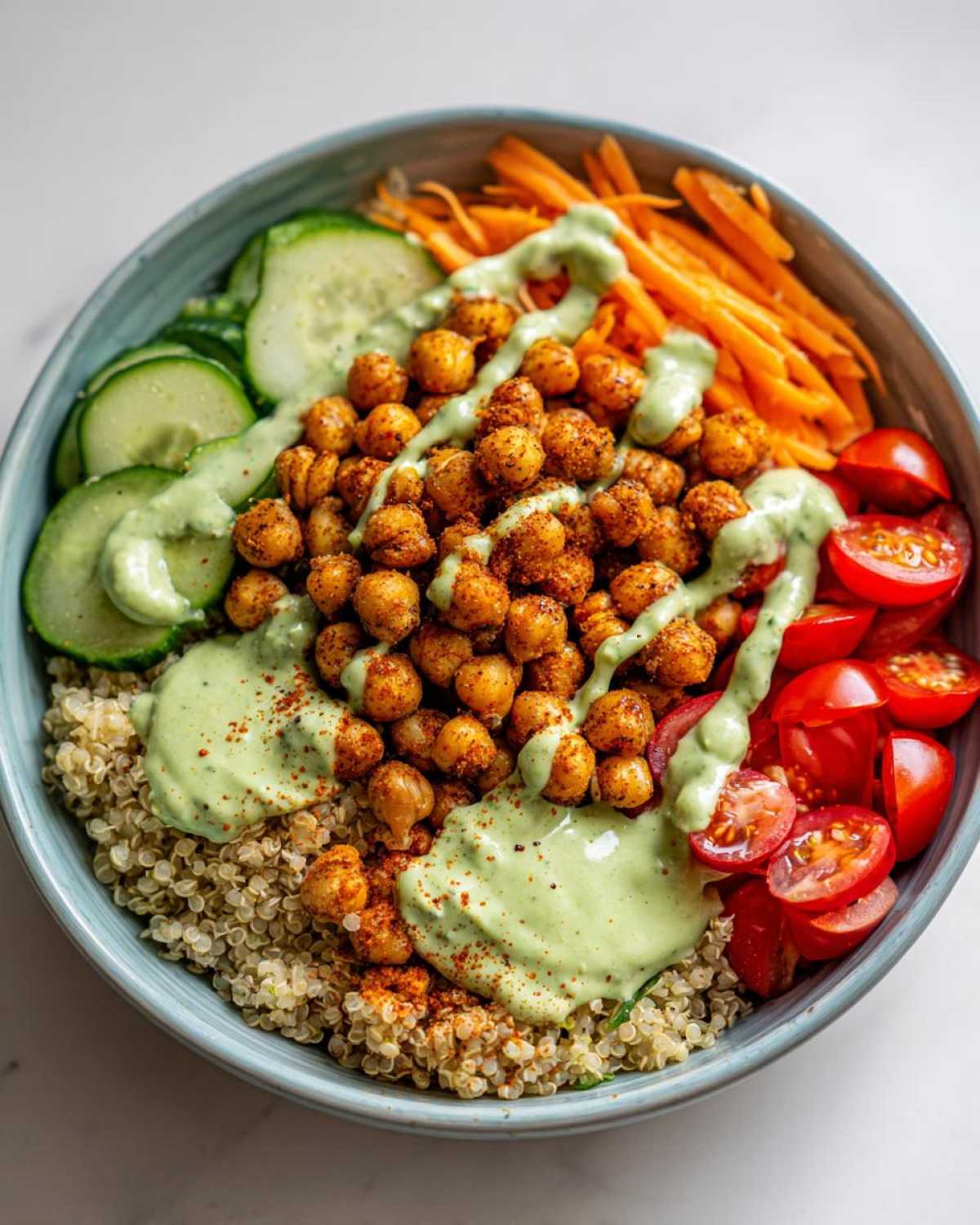 Close-up of a vibrant Chickpea Grain Bowl with avocado lemon tahini dressing over quinoa, roasted chickpeas, cucumbers, carrots, and tomatoes.