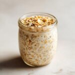 Close-up of creamy Overnight Oats soaking in milk with chia seeds, topped with dry rolled oats in a glass jar.