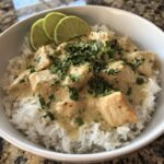 A close-up of a white bowl filled with white rice, topped with creamy Coconut Chicken Bowl pieces and fresh cilantro, garnished with lime slices.