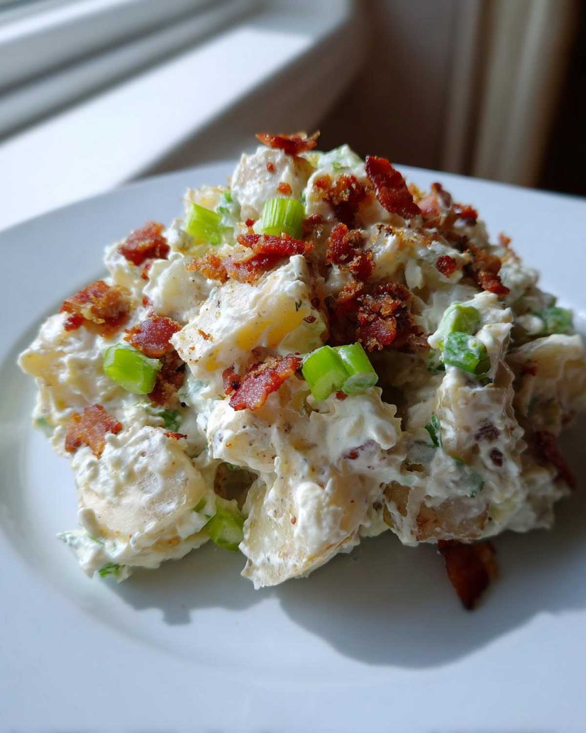 A scoop of creamy Baked Potato Salad topped with crispy bacon bits and sliced green onions on a white plate.