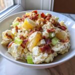 A close-up of creamy Baked Potato Salad topped with crispy bacon bits, chopped celery, and chunks of hard-boiled egg.