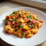 A close-up of creamy, orange-coated Bang Bang Salmon Salad topped with fresh cilantro and diced red onion on a white plate.