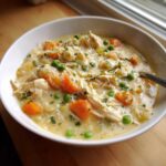 A close-up of a creamy bowl of Chicken Pot Pie Soup, featuring shredded chicken, carrots, and peas.