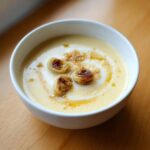 Close-up of creamy Garlic Soup with Roasted Bulb halves, cream swirl, and crispy topping.