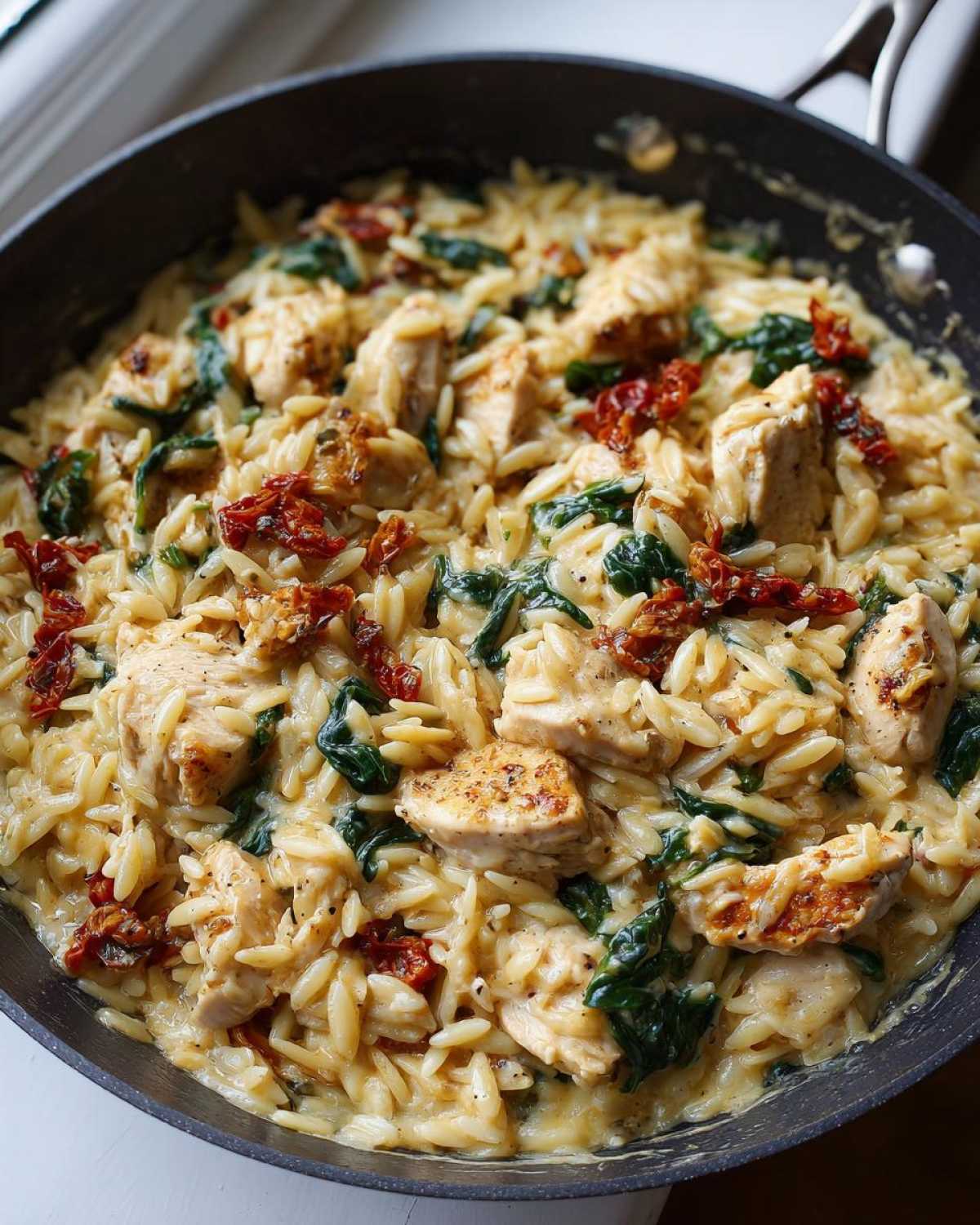 Close-up of creamy Marry Me Chicken Orzo mixed with spinach and sun-dried tomatoes in a black skillet.