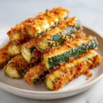 A close-up stack of golden brown Crispy Baked Parmesan Zucchini sticks piled on a light plate.