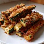 A close-up of a pile of golden brown Crispy Baked Parmesan Zucchini sticks on a white plate.