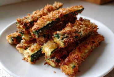 A close-up of a pile of golden brown Crispy Baked Parmesan Zucchini sticks on a white plate.