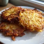 Close-up of several golden-brown, crispy German Potato Pancakes stacked on a white plate, illuminated by natural light.