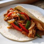 A single serving of Crockpot Chicken Fajitas filling with sliced peppers and onions served in a warm flour tortilla on a white plate.